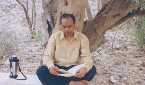 Bhaiyaji seated beneath a tree, looking over pages of the manuscript